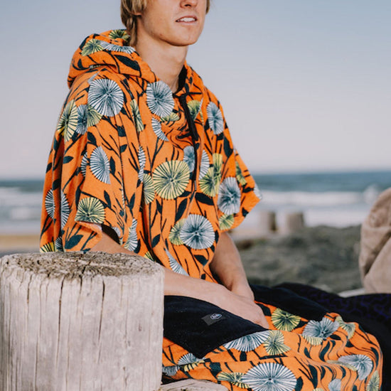 Homme portant un poncho de surf ALL-IN coupe V-Shape modèle Dandelion orange à motifs fleurs sur une plage.
