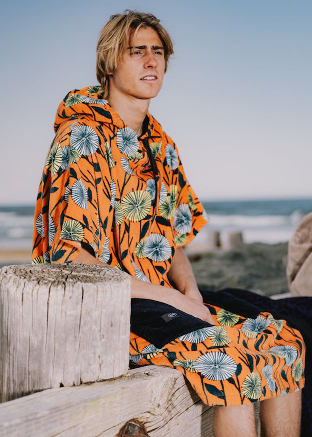 Homme portant un poncho de surf ALL-IN coupe V-Shape modèle Dandelion orange à motifs fleurs sur une plage.